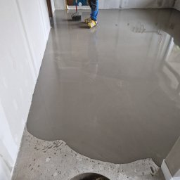 BUD-MAT - Wylewka samopoziomująca na podłodze w trakcie rozprowadzania wałkiem kolczastym, widoczne nogi pracownika w obuwiu ochronnym.