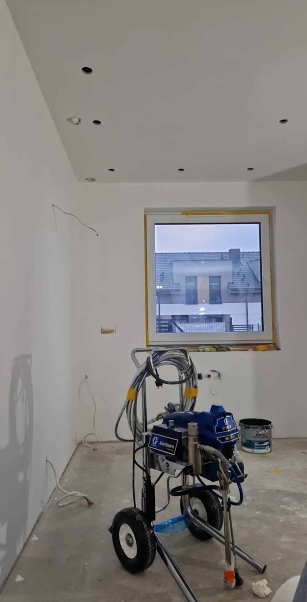 Wnętrze w trakcie malowania natryskowego: białe ściany z widocznymi otworami na oświetlenie, okno zabezpieczone taśmą malarską, agregat malarski na pierwszym planie oraz wiadro z farbą w tle.