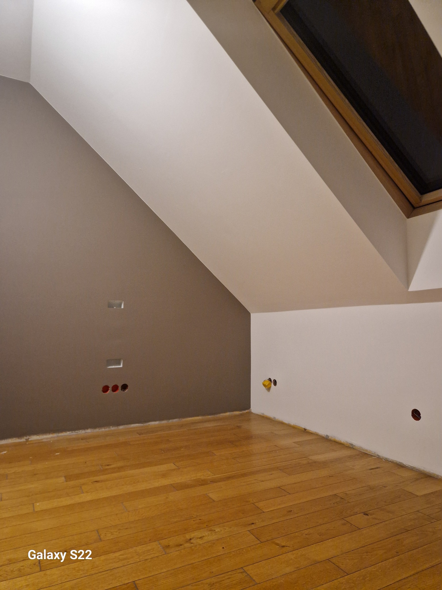 Poddasze po remoncie: drewniana podłoga, ściana pomalowana na dwa kolory (brązowy i biały), widoczne gniazdka elektryczne i otwory na kable, okno dachowe.