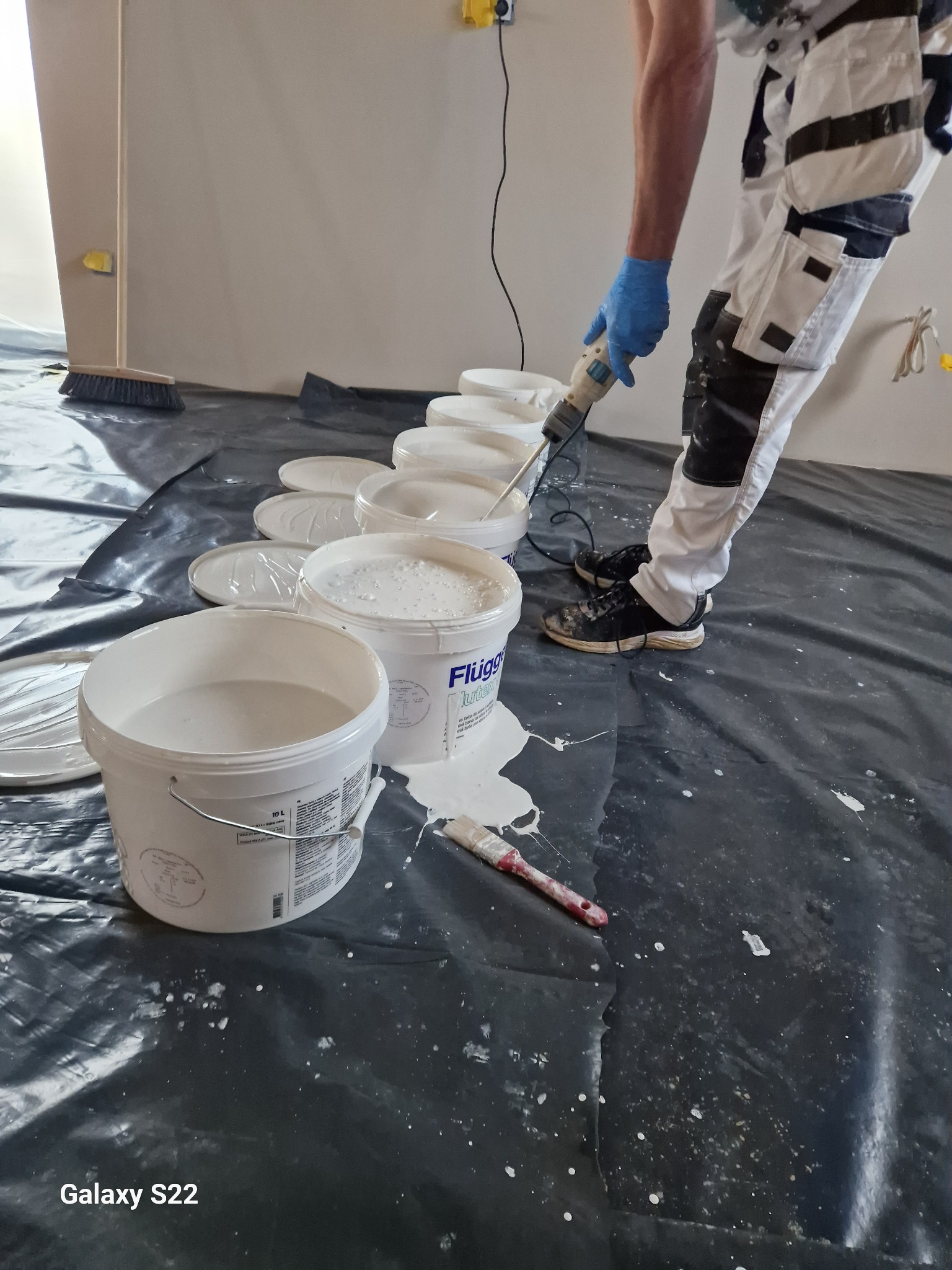 Mężczyzna w roboczym ubraniu miesza farbę Flugo w wiadrze za pomocą mieszadła elektrycznego, widoczne otwarte pojemniki z farbą i pędzel na folii ochronnej.