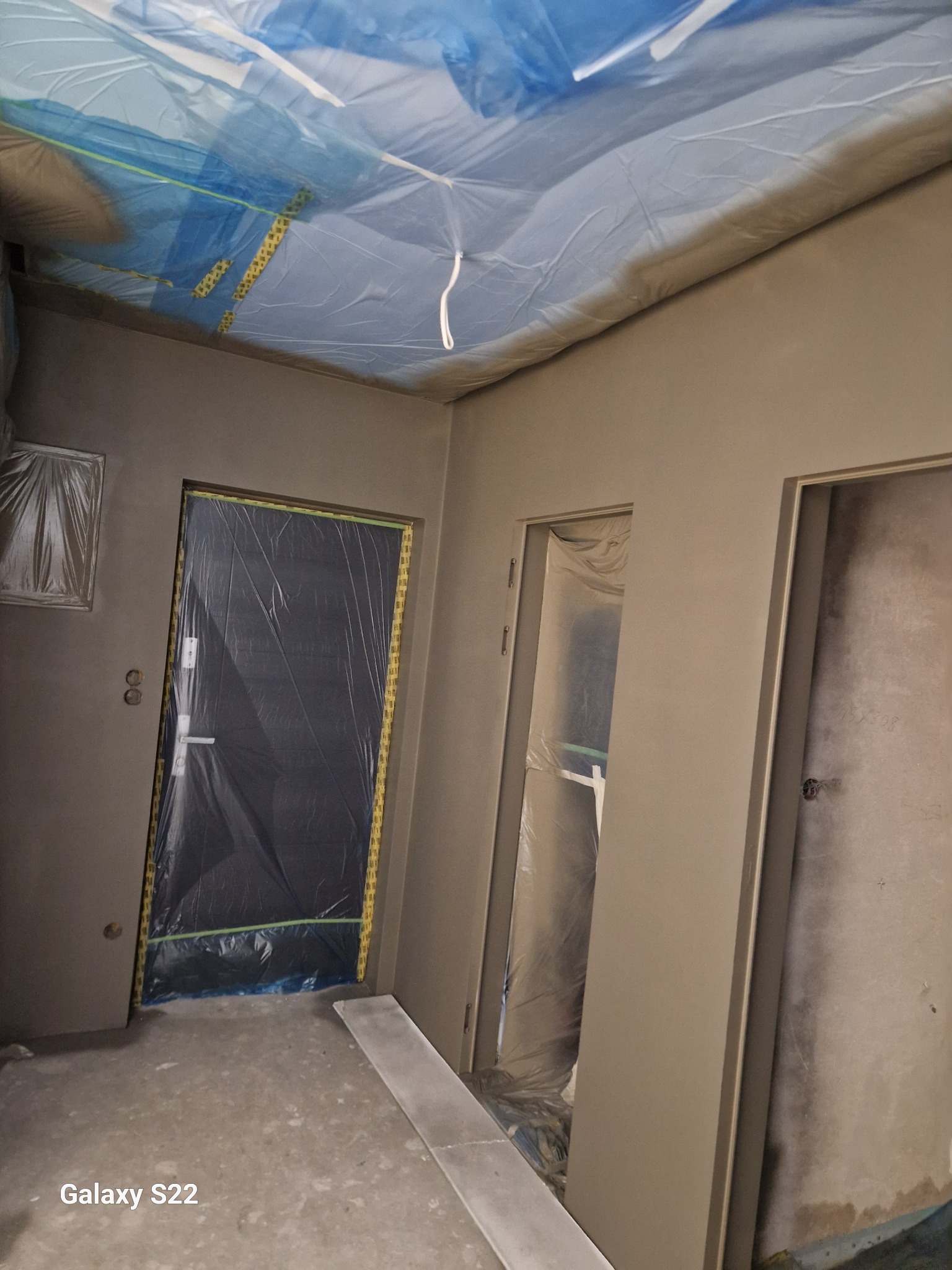 Przygotowanie do malowania ścian w pomieszczeniu: ściany pokryte świeżą gładzią, drzwi zabezpieczone folią malarską, sufit osłonięty niebieską folią ochronną.