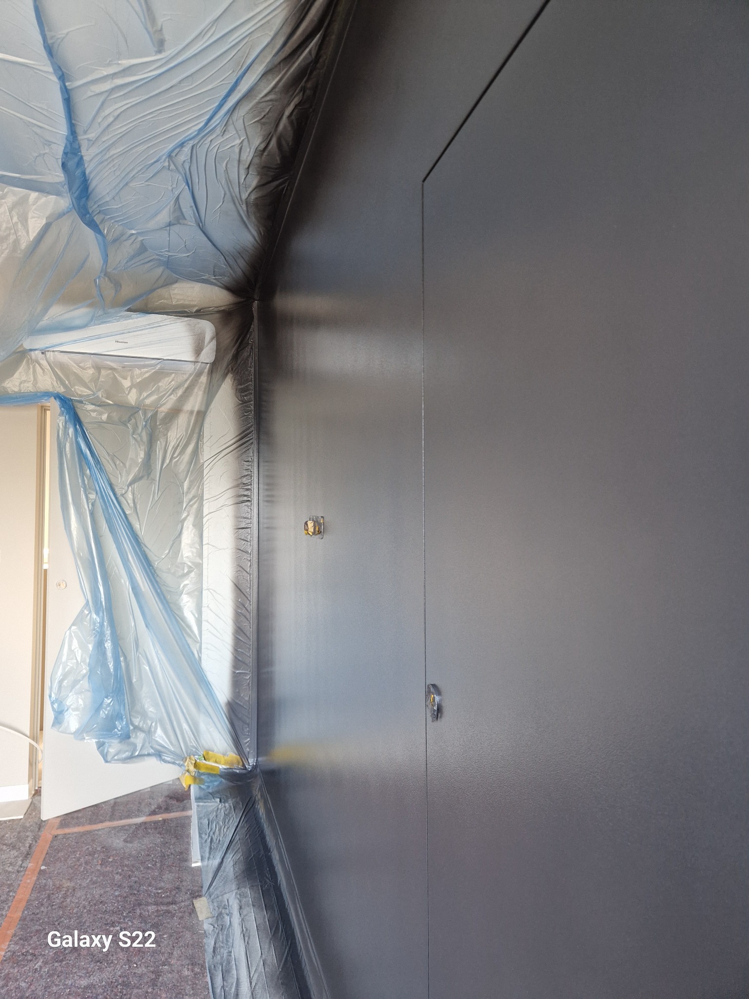 Świeżo pomalowana, ciemnoszara ściana z widocznymi zabezpieczeniami folią malarską na klimatyzatorze i framudze drzwi, widoczne gniazdko elektryczne, wykonane z bliskiej odległości.