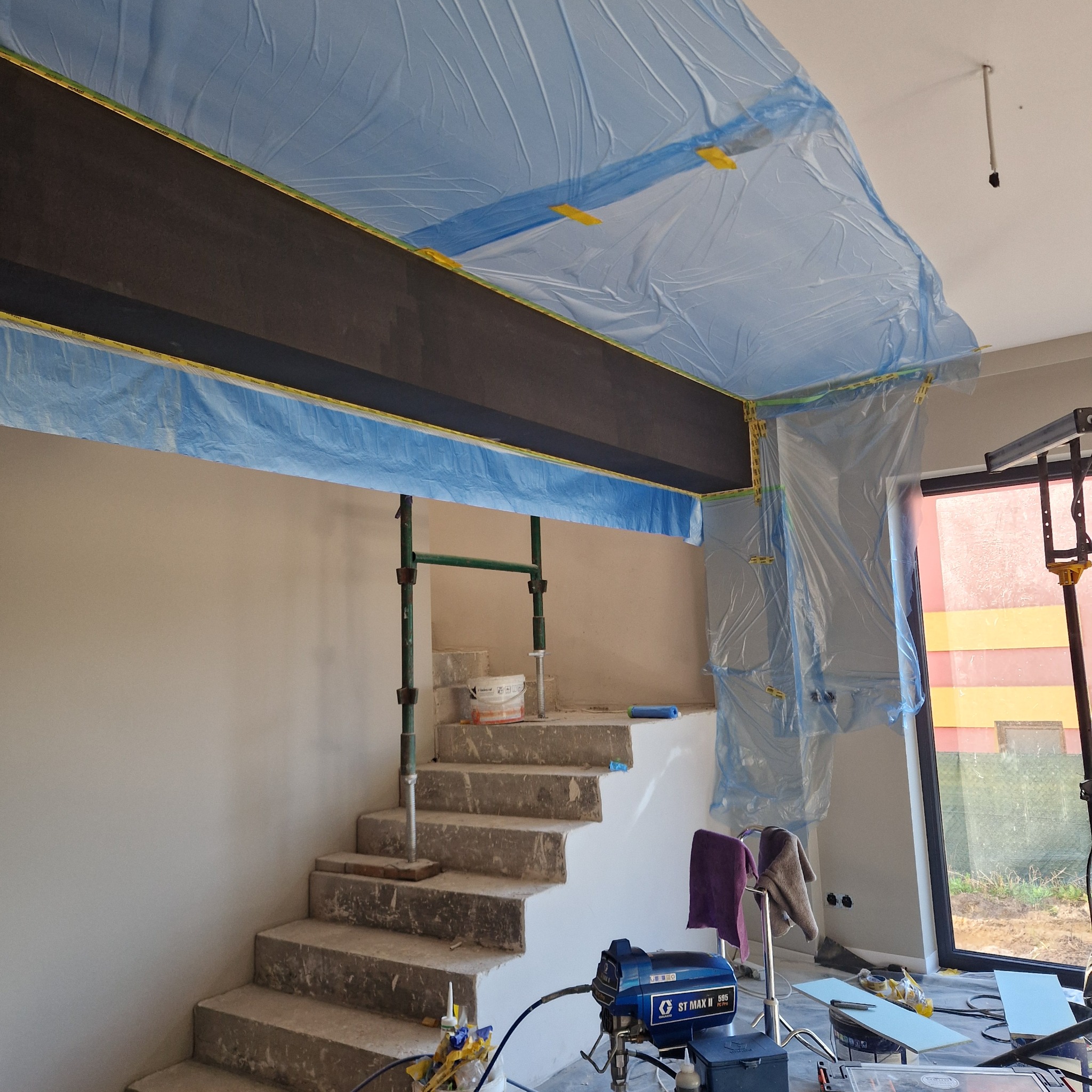 Przygotowanie do malowania wnętrza: schody zabezpieczone folią, ściany zagruntowane, widoczny agregat malarski Graco ST MAX II 595 oraz drabina.