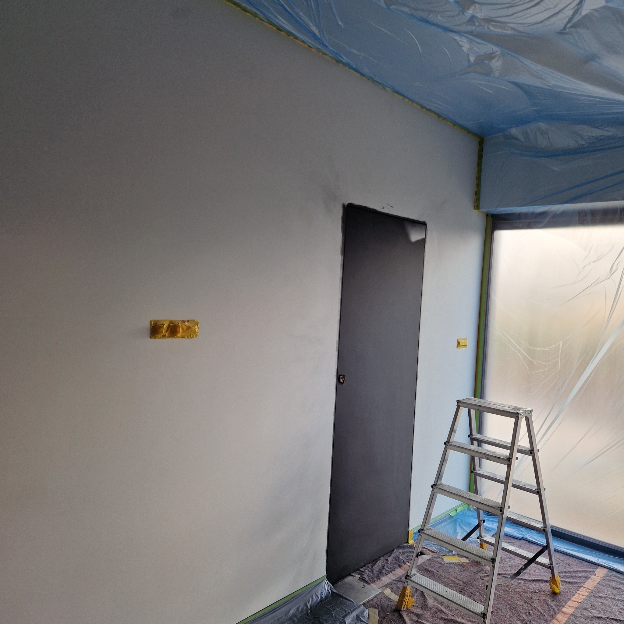 Przygotowanie do malowania: ściana po szpachlowaniu, zabezpieczona folią i taśmą malarską, drabina aluminiowa z zabezpieczonymi nogami.