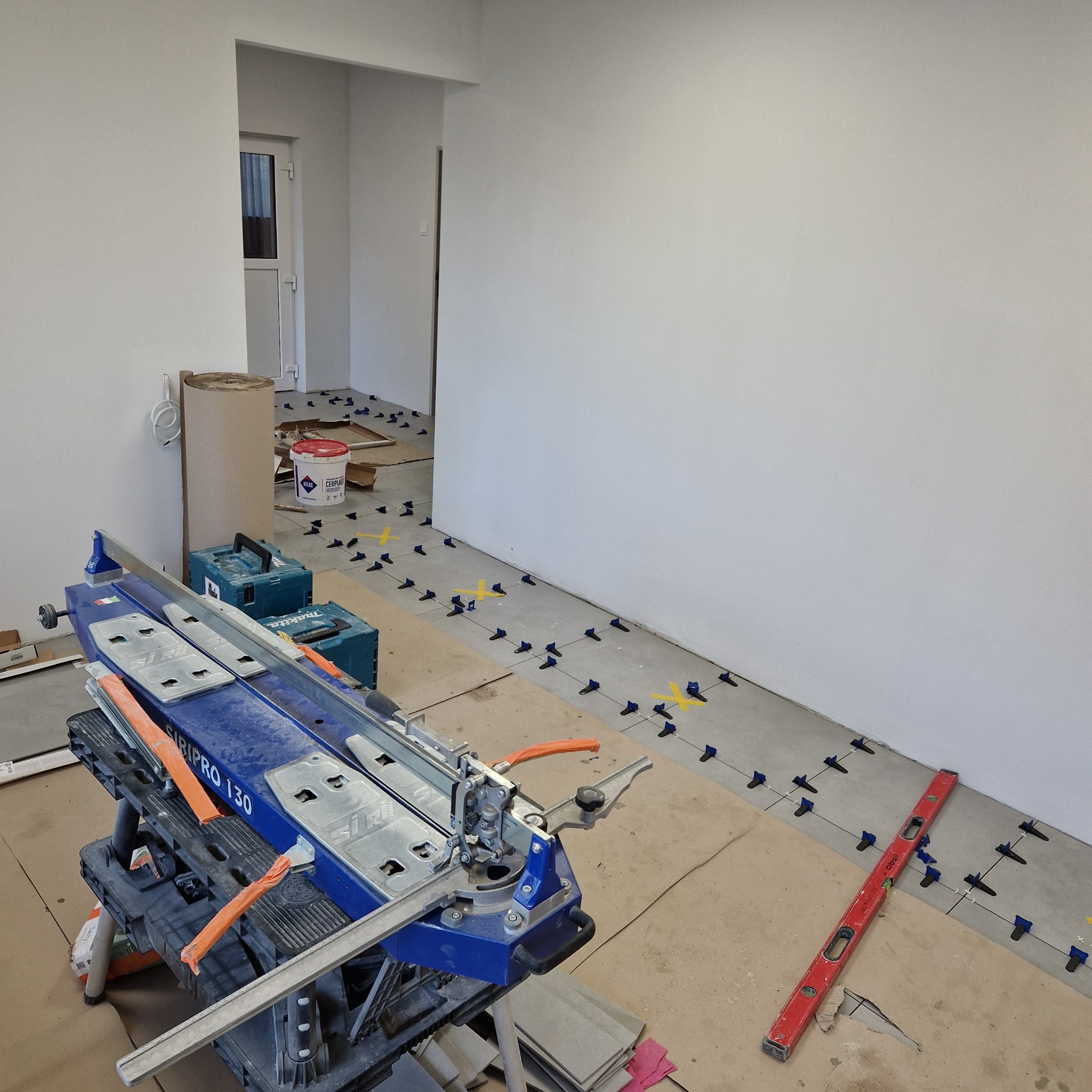 Układanie płytek podłogowych: widoczny niebieski przecinak do płytek Siripro 130, poziomica, wiadro z klejem, krzyżyki dystansowe i fragment ułożonej podłogi w jasnym pomieszczeniu.