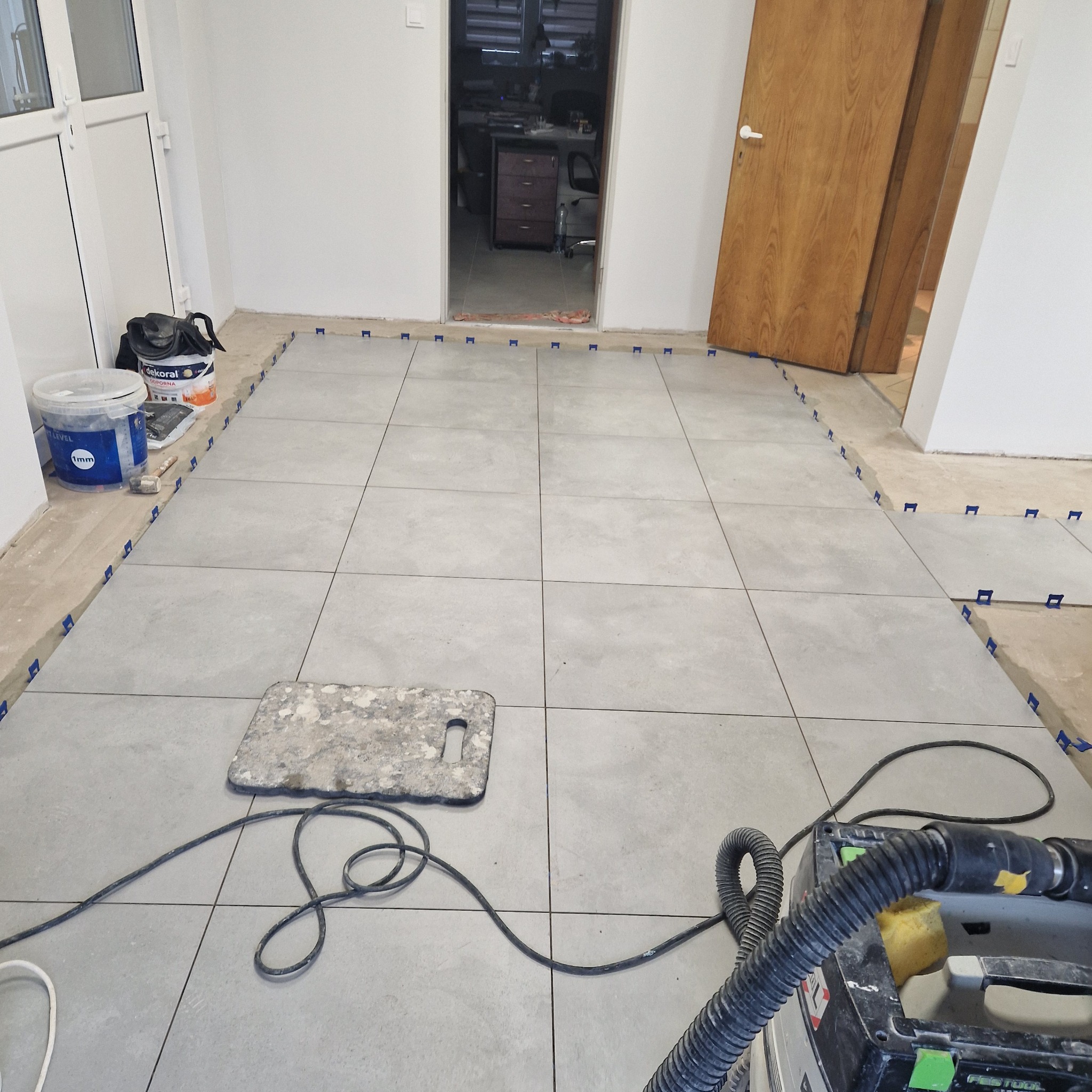 Układanie szarych płytek podłogowych w pomieszczeniu, widoczne niebieskie kliny poziomujące, gąbka, odkurzacz przemysłowy Festool oraz wiadro z farbą Dekoral obok białych drzwi.