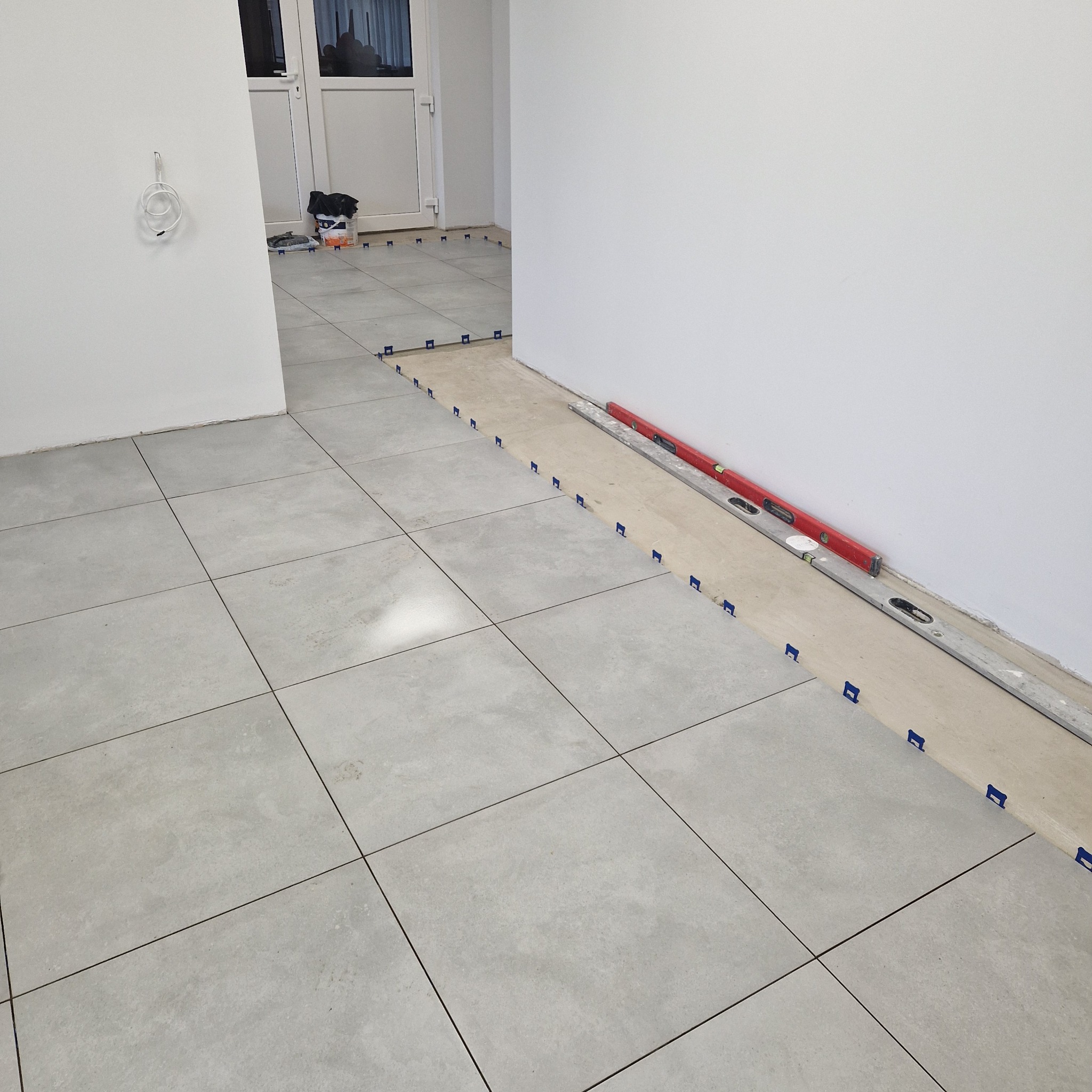 Szare płytki podłogowe ułożone w kwadraty z plastikowymi klipsami dystansowymi, poziomice przy ścianie, widok na podłogę w trakcie układania.