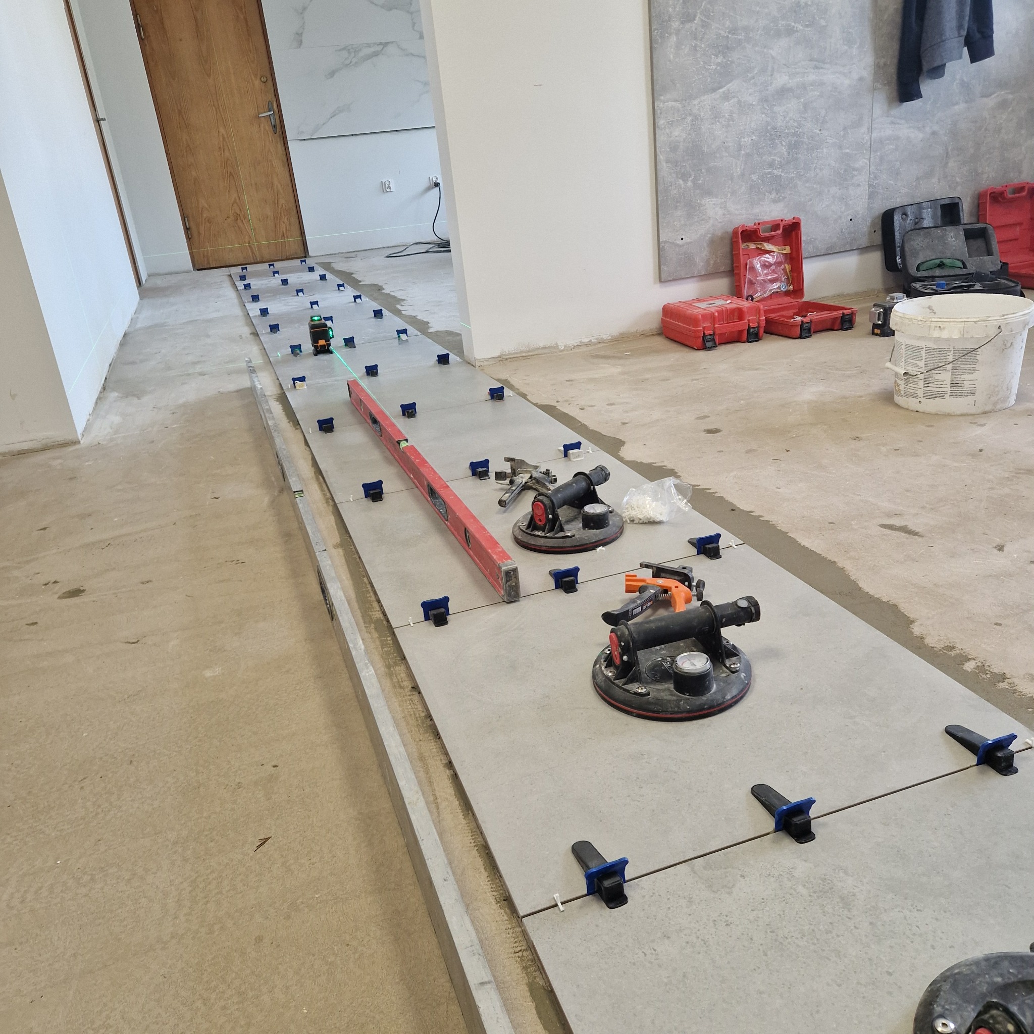 Układanie szarych płytek podłogowych z użyciem poziomicy laserowej, przyssawek i klipsów poziomujących; widoczny także wiadro i skrzynki z narzędziami.