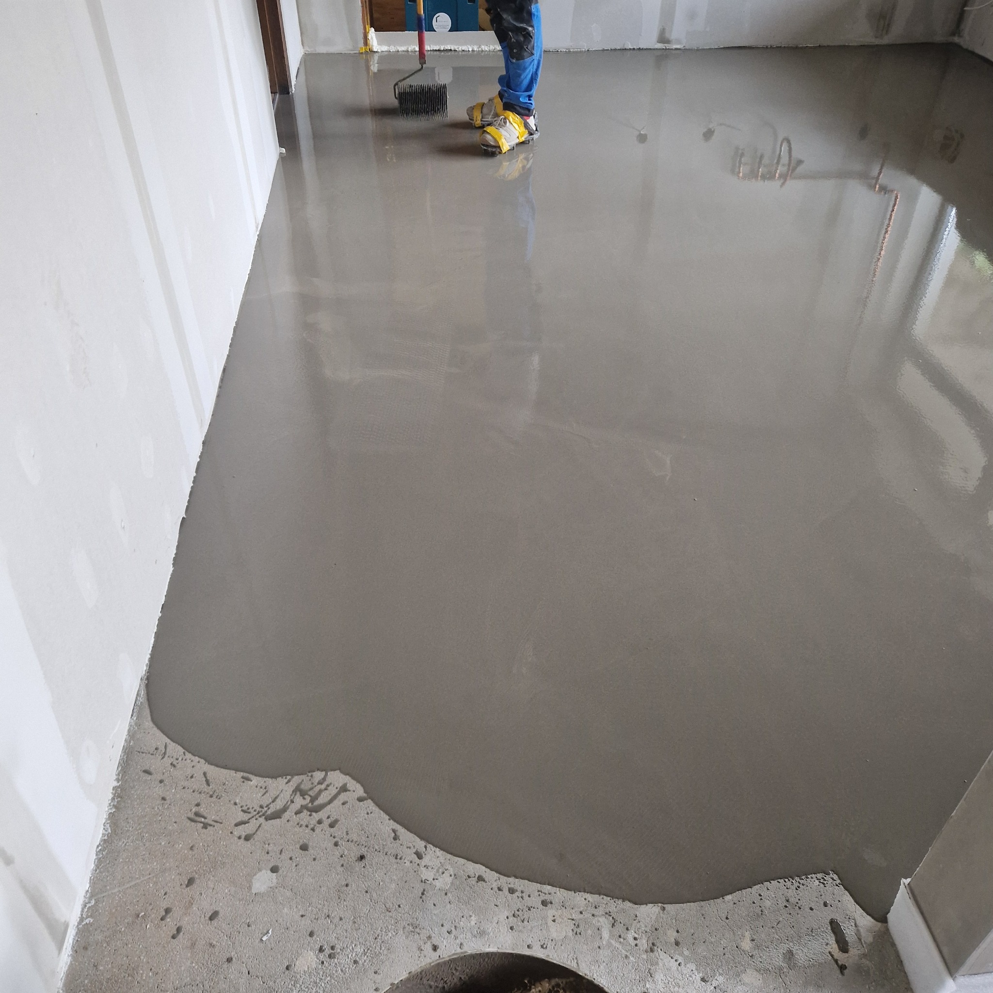 Wylewka samopoziomująca na podłodze w trakcie rozprowadzania wałkiem kolczastym, widoczne nogi pracownika w obuwiu ochronnym.
