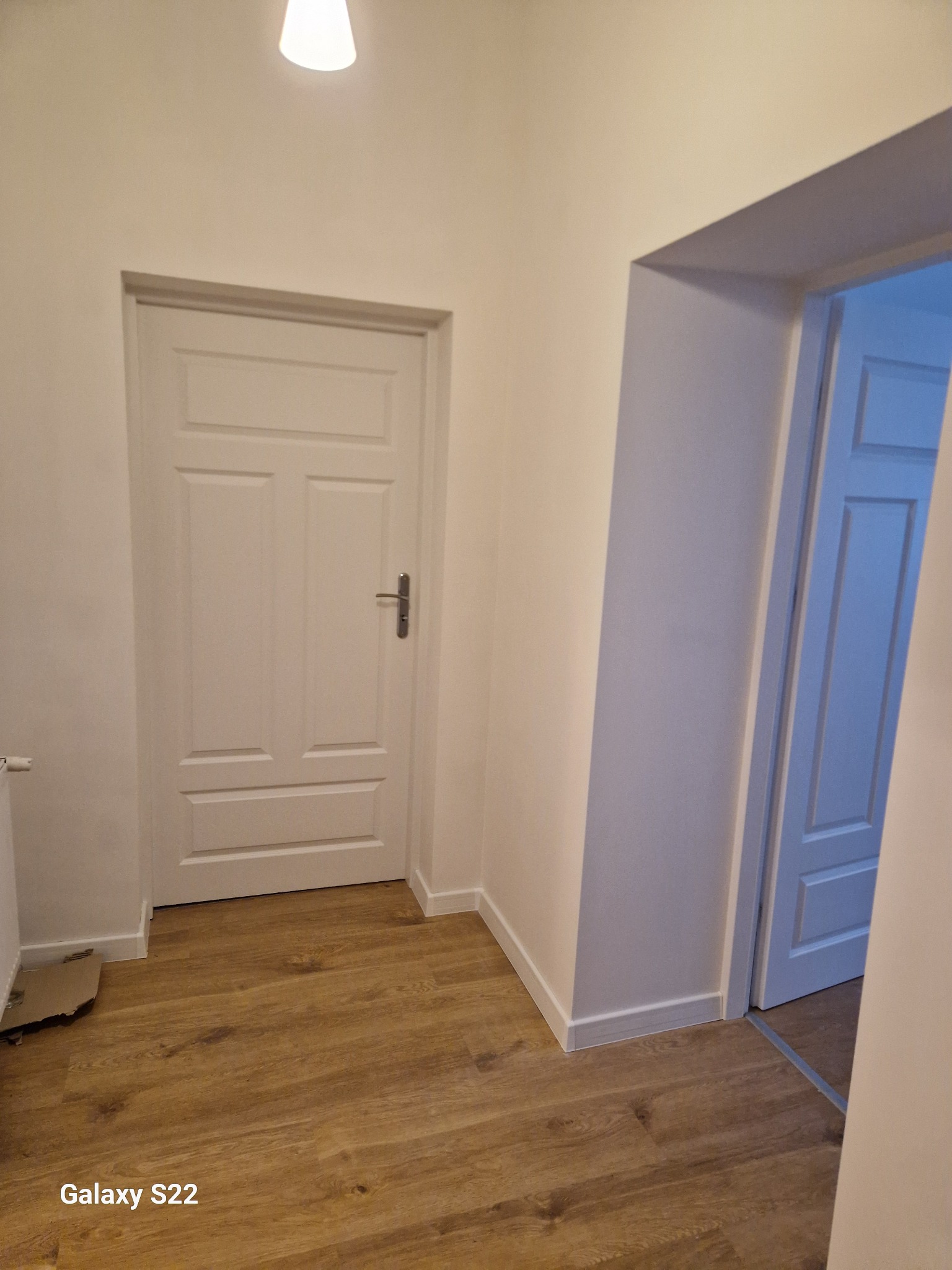 Korytarz po remoncie z panelami podłogowymi w odcieniach brązu i dwiema białymi, odnowionymi drzwiami; jedna prowadzi do pomalowanego na niebiesko pomieszczenia. Widoczne listwy przypodłogowe.