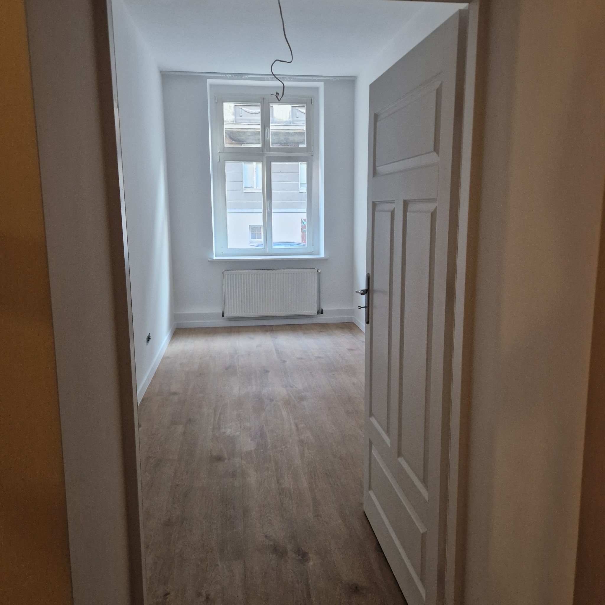 Pusty pokój po remoncie z drewnianą podłogą, białymi ścianami i grzejnikiem pod oknem, widok z korytarza przez otwarte białe drzwi.