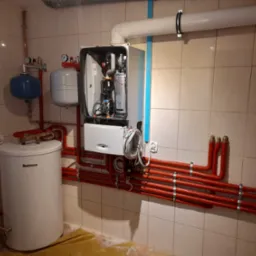 Nowoczesny piec gazowy Junkers z otwartą obudową, zbiornik Buderus, instalacja z czerwonych rur w pomieszczeniu wyłożonym kafelkami.