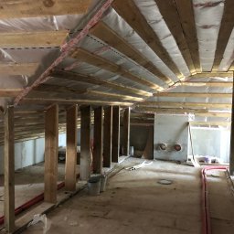 DAVLATBUD - Poddasze w trakcie adaptacji: drewniane belki stropowe z folią izolacyjną, pionowe słupy konstrukcyjne, instalacje rurowe i surowa podłoga w nieukończonym pomieszczeniu.