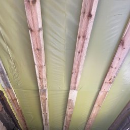 DAVLATBUD - Widok z dołu na drewniane belki stropowe zabezpieczone żółtą folią izolacyjną, widoczne ślady wilgoci na ścianach.