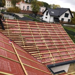 DAVLATBUD - Pracownik w czapce montuje drewniane łaty na czerwonej membranie dachowej, w tle domy jednorodzinne i jesienne drzewa.