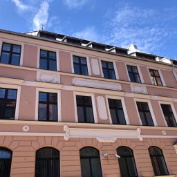 DAVLATBUD - Fasada odnowionego, trzypiętrowego budynku z różową elewacją, czarnymi oknami i ozdobnymi elementami architektonicznymi pod błękitnym niebem.