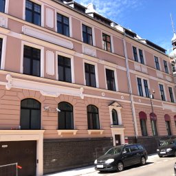 DAVLATBUD - Odnowiona, różowa elewacja kamienicy z czarnymi oknami i białymi detalami architektonicznymi, z wieżą w rusztowaniach w tle i zaparkowanymi samochodami na pierwszym planie.