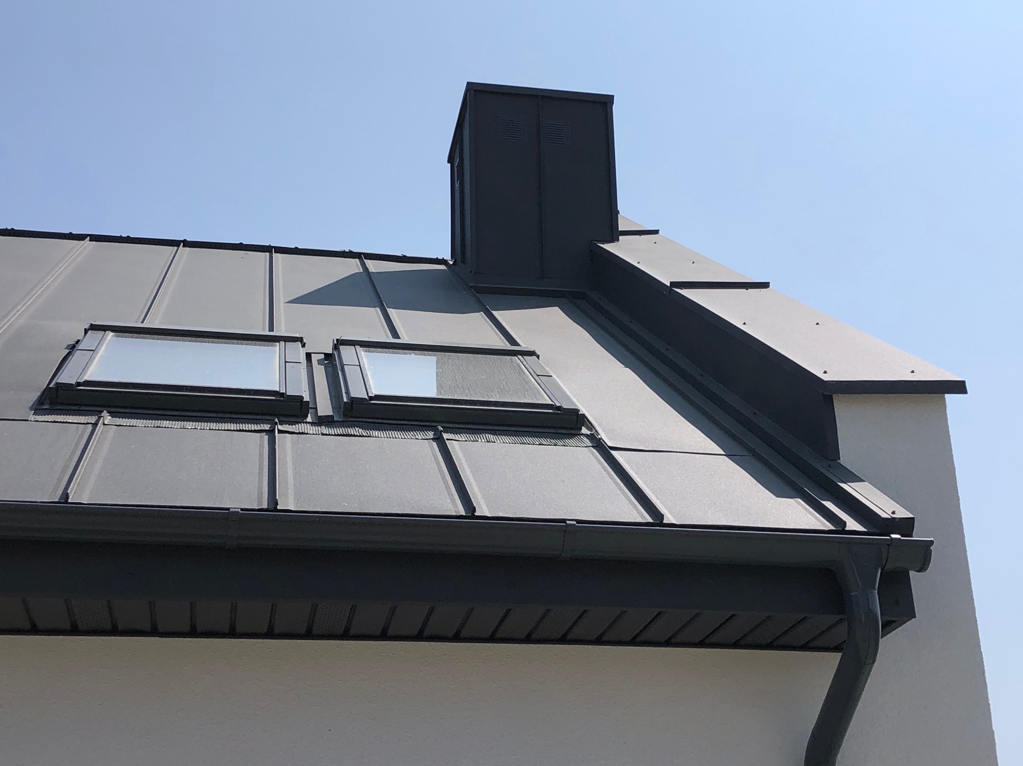 Szary blaszany dach z dwoma oknami dachowymi i kominem wentylacyjnym na tle błękitnego nieba, widok z dołu, fragment ściany budynku.