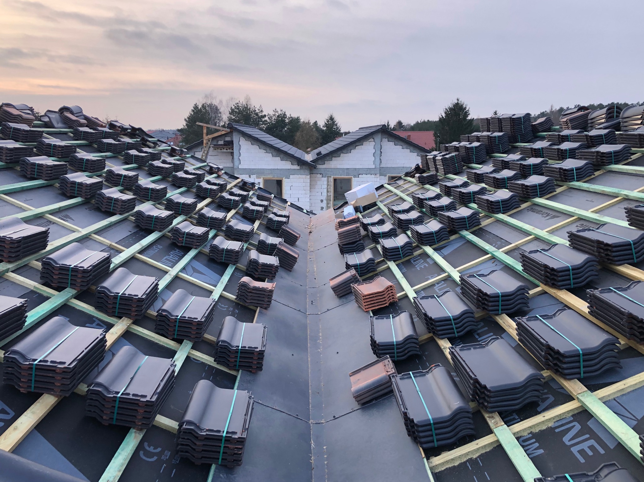 Stosy ciemnych dachówek ceramicznych z zielonymi taśmami na drewnianej konstrukcji dachu w trakcie budowy, widok z góry na tle surowego stanu ścian budynku.