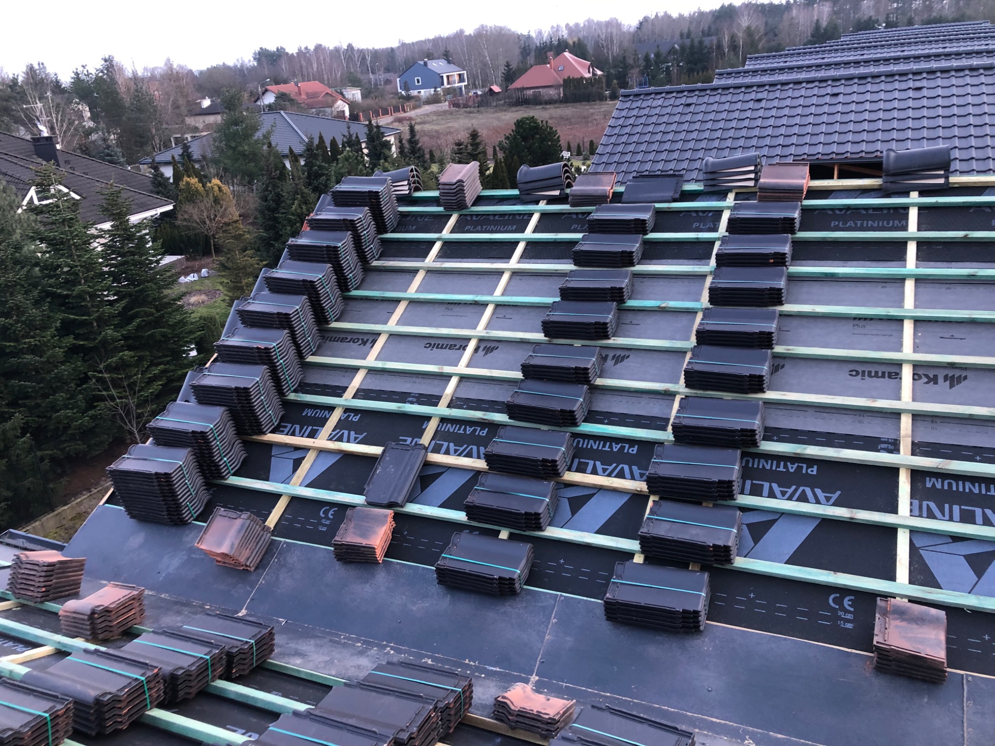 Układanie czarnej dachówki ceramicznej na nowym dachu, widoczne łaty, membrana dachowa z oznaczeniami producenta oraz sterty dachówek przygotowane do montażu.