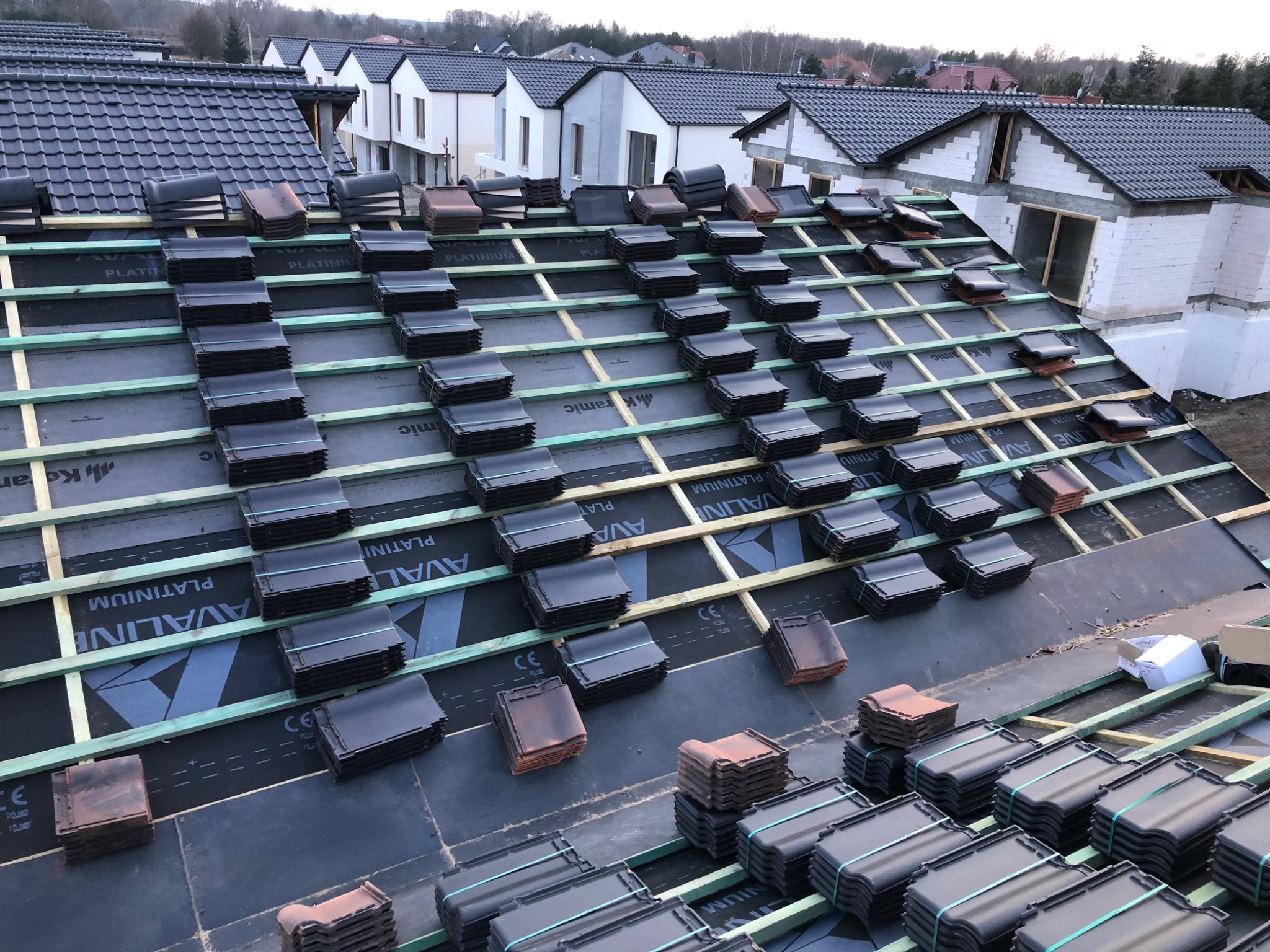 Przygotowanie do układania dachówki ceramicznej na nowym dachu, widoczne łaty, membrana dachowa i ułożone w rzędach paczki z czarną dachówką oraz pojedyncze brązowe dachówki, w tle domy w stanie...