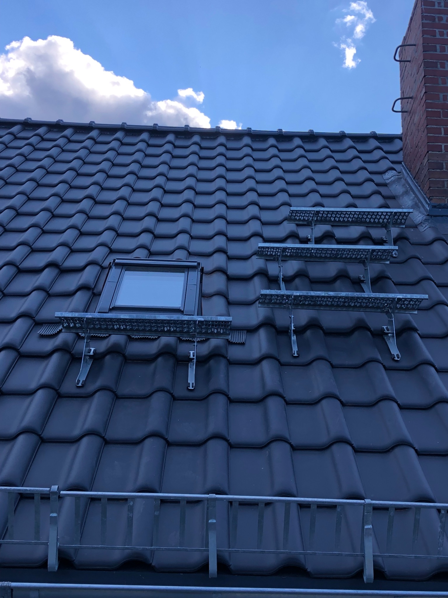 Ciemna dachówka ceramiczna z zamontowanym oknem dachowym i metalowymi pomostami roboczymi, widok na błękitne niebo z chmurami.