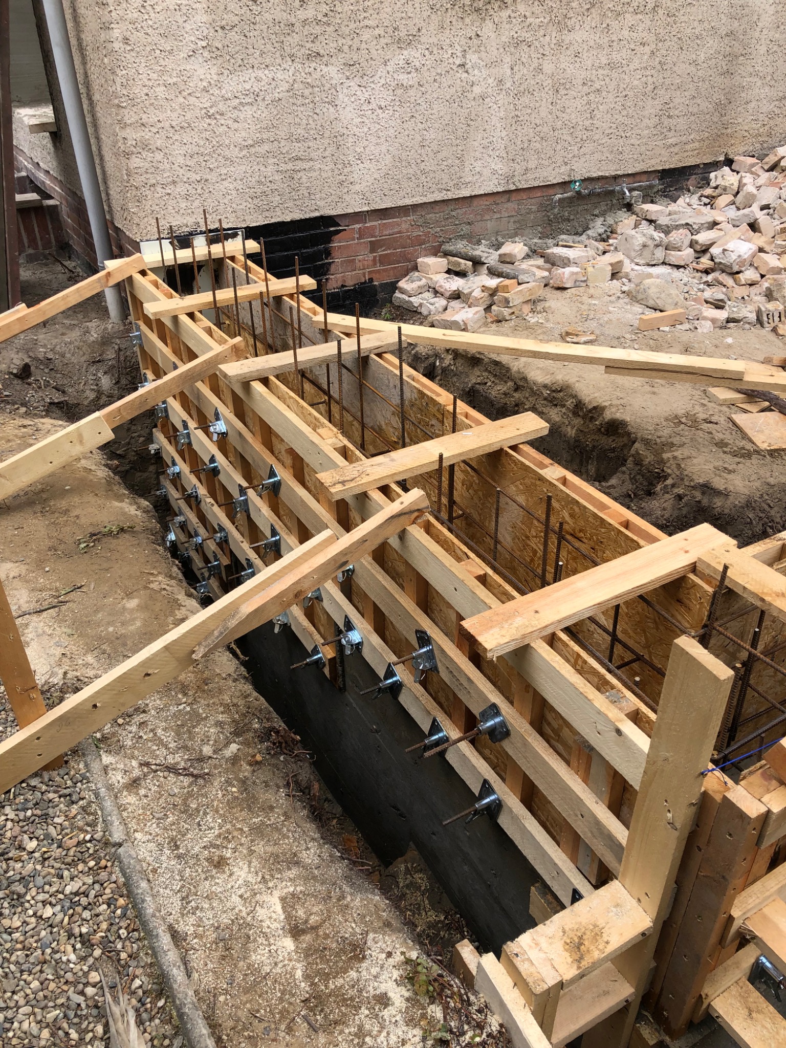Szczegółowe ujęcie drewnianego szalunku zbrojeniowego fundamentu, widoczne metalowe elementy mocujące, pręty zbrojeniowe, wykop ziemny oraz fragment ściany budynku z widocznymi uszkodzeniami.