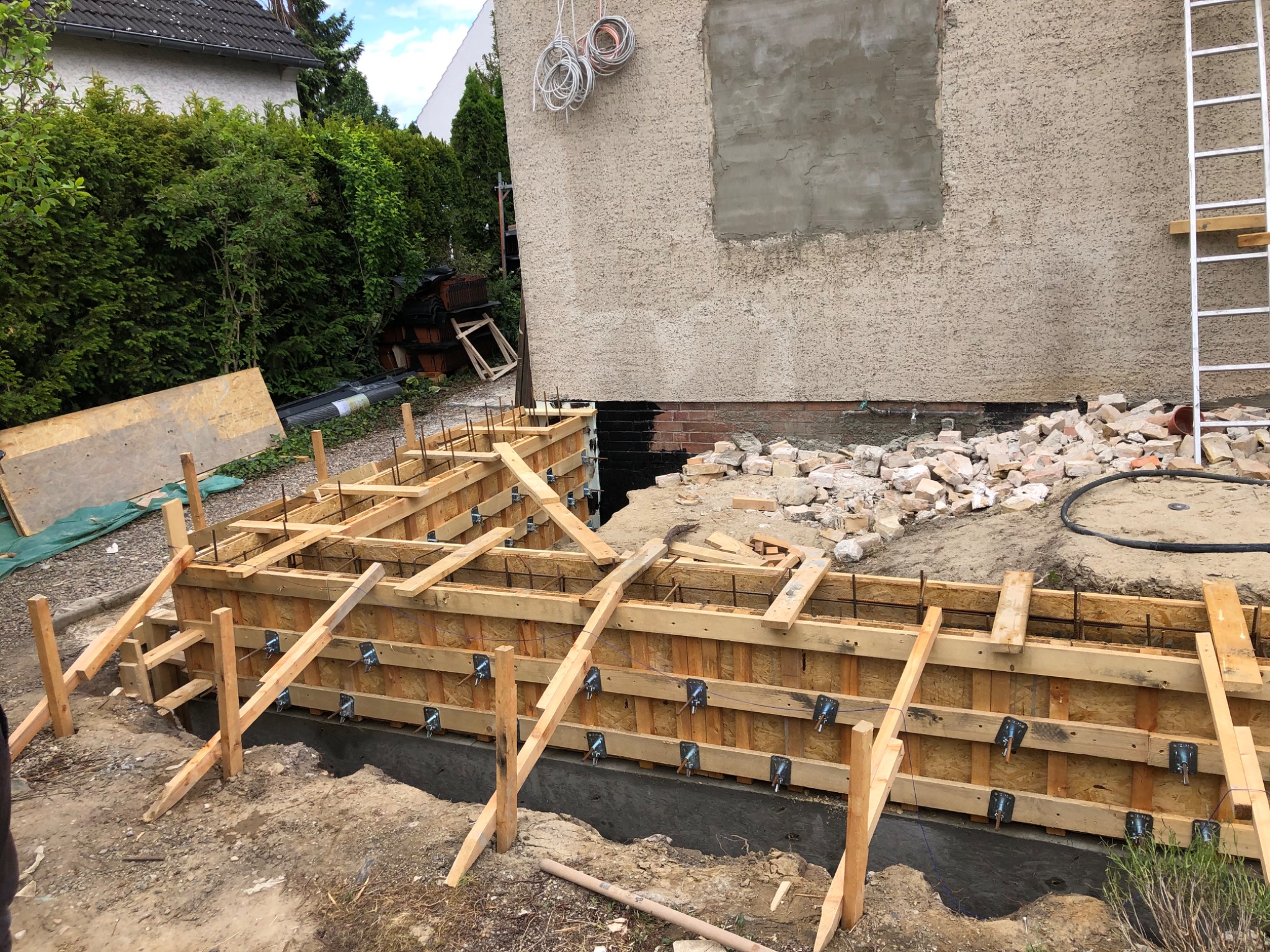 Szlifowanie fundamentów domu jednorodzinnego, widoczne drewniane szalunki z metalowymi ściągami, zarys muru pomalowany na czarno, gruz i drabina oparta o ścianę budynku.