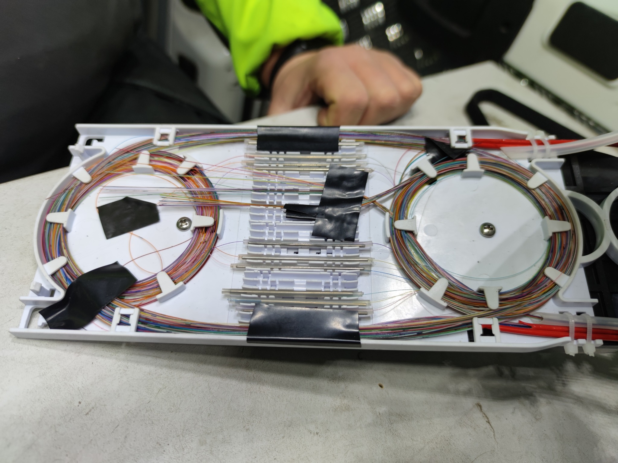 Otwarta skrzynka światłowodowa z precyzyjnie ułożonymi kablami światłowodowymi w różnych kolorach, zabezpieczonymi czarną taśmą, widoczna dłoń pracownika w odblaskowej kurtce w tle.