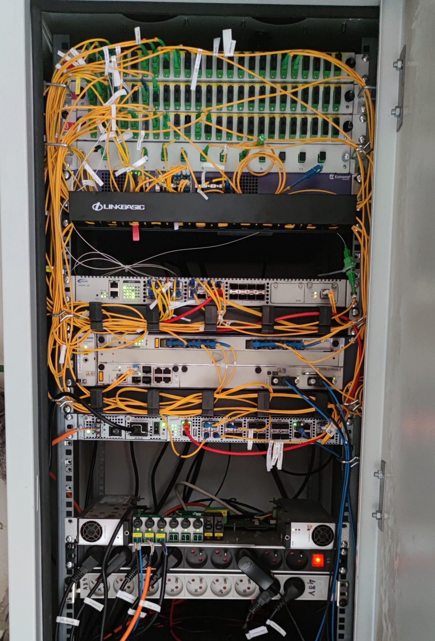 Szafa telekomunikacyjna z zainstalowanymi urządzeniami sieciowymi, licznymi kablami światłowodowymi oznaczonymi etykietami, zasilaczami i listwami zasilającymi, widoczne marki LINKBASIC i DASAN