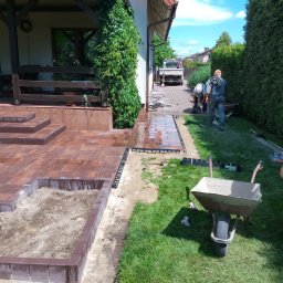 DiP BRUK Damian Szymański - Montaż obrzeży trawnikowych przy nowej, brązowej kostce brukowej na posesji; w tle pracownicy układają kostkę na podjeździe, a przy budynku pnie się zielony bluszcz.