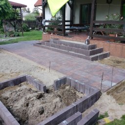 DiP BRUK Damian Szymański - Układanie ścieżki z kostki brukowej w ogrodzie, widoczne obrzeża z kostki, częściowo ułożona nawierzchnia i przygotowany wykop pod kolejne elementy, w tle dom z tarasem.