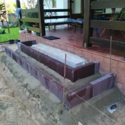 Montaż obrzeży z ciemnej kostki brukowej przy tarasie domu, widoczne sznurki wyznaczające linię, metalowe pręty, piasek i drewniany kołek z nawiniętym sznurkiem.