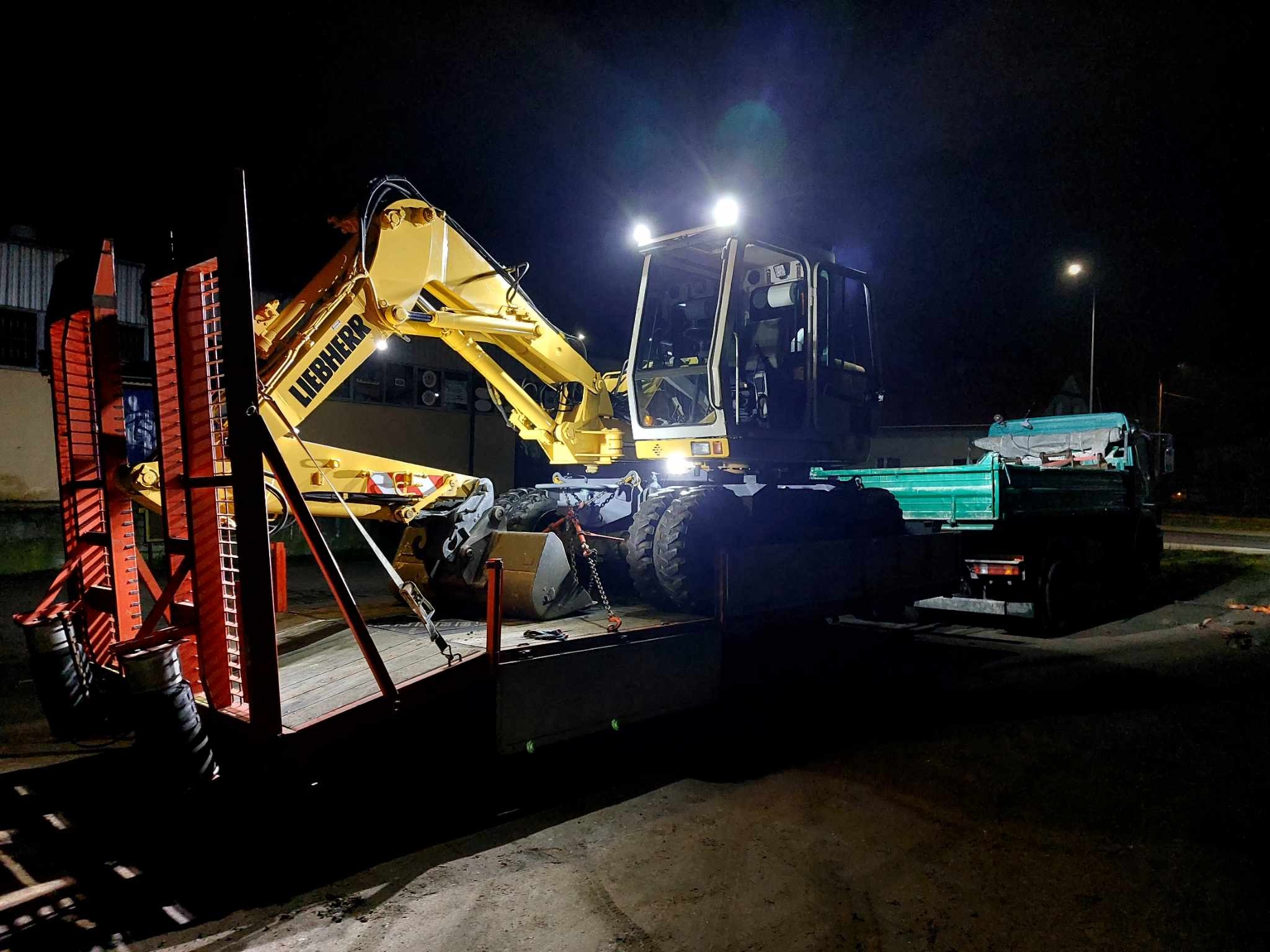 Żółta koparka Liebherr z włączonymi światłami, zabezpieczona łańcuchami na platformie transportowej ciężarówki, widoczna w nocy na tle budynku.