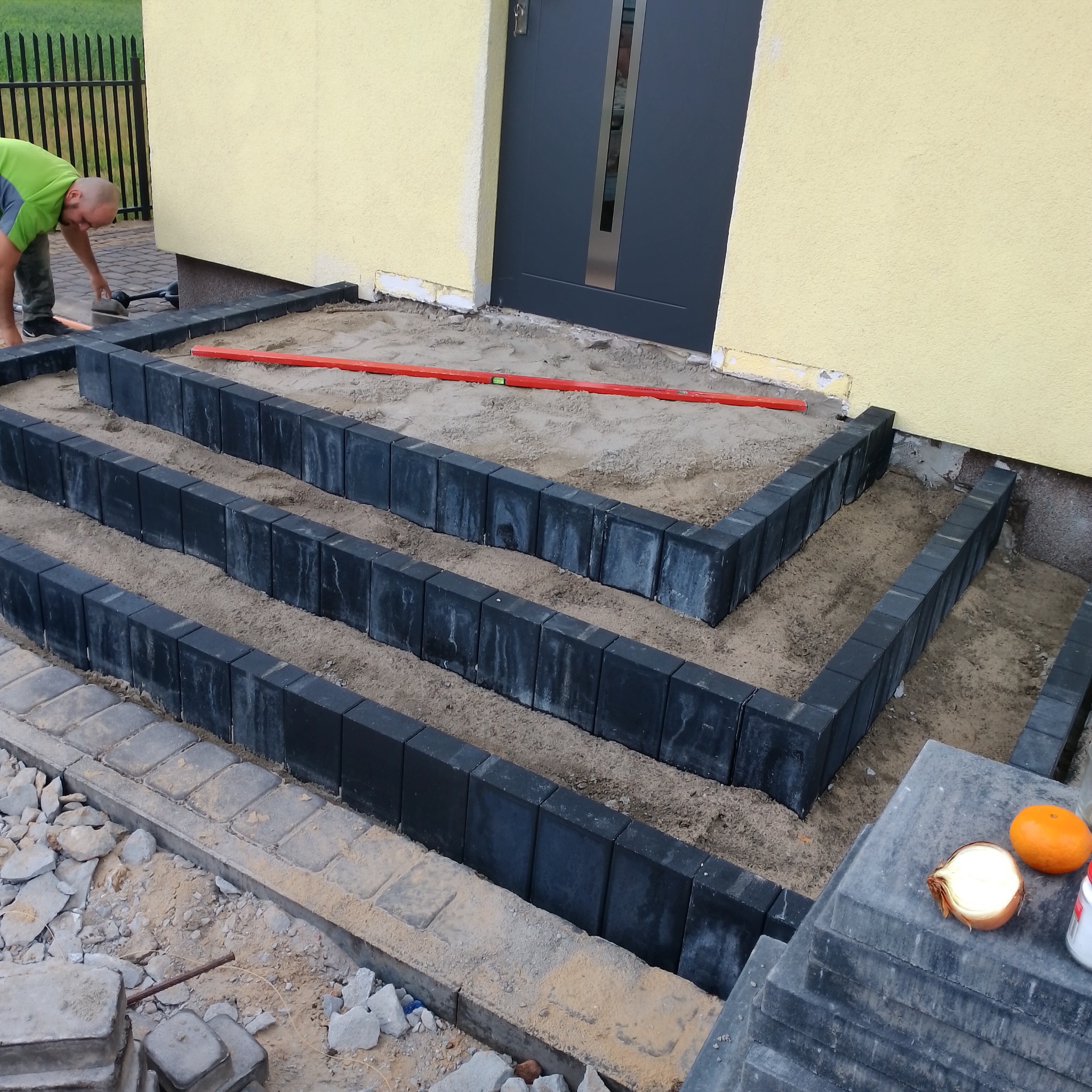 Budowa schodów wejściowych z czarnej kostki brukowej, widoczny pracownik z poziomnicą, przygotowane podłoże z piasku, cebula i mandarynka na stosie kostki obok.