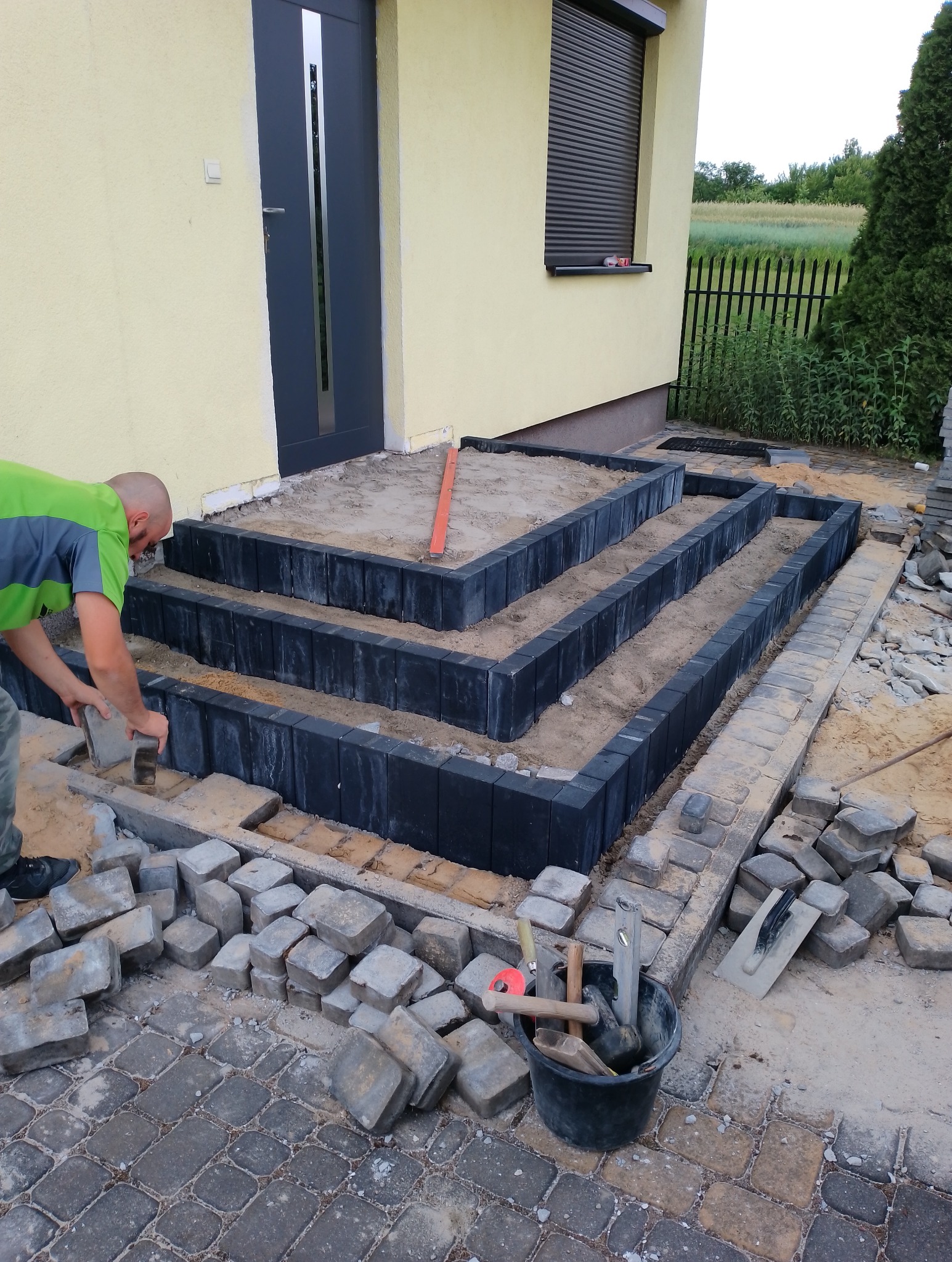 Pracownik w zielonej koszulce układa szare kostki brukowe na schodach wejściowych do domu z żółtą elewacją, widoczne narzędzia w czarnym wiadrze i poziomica.