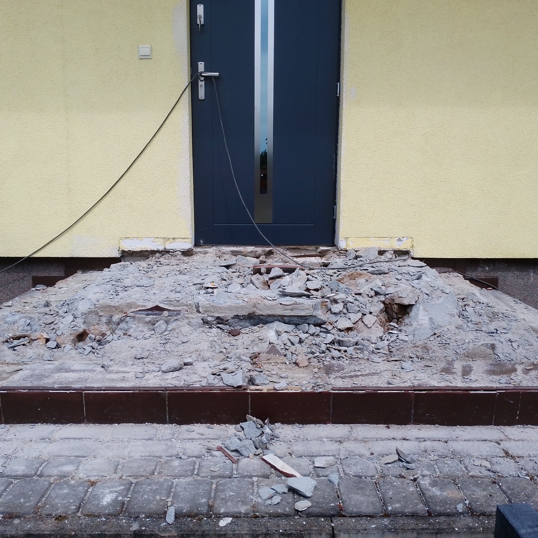 Demontaż schodów wejściowych z kostki przed budynkiem o żółtej elewacji i ciemnych drzwiach, widoczne gruz i pozostałości po rozbiórce.