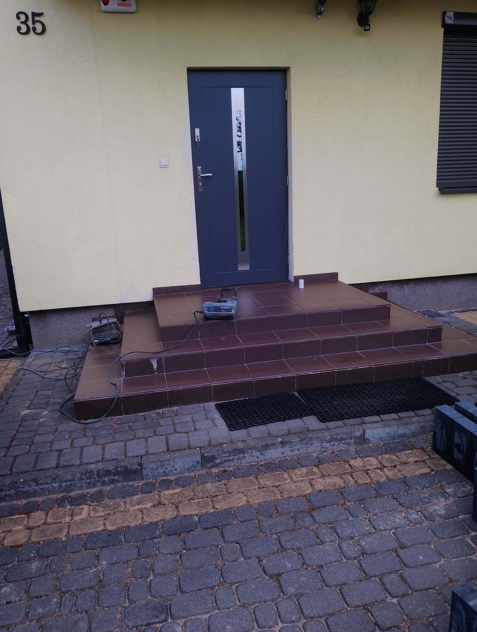 Wykończone schody wejściowe do domu z ciemnobrązowych płytek, na dole ułożona kostka brukowa w dwóch odcieniach szarości i żółci, widoczny sprzęt budowlany na schodach i zwinięty kabel obok.