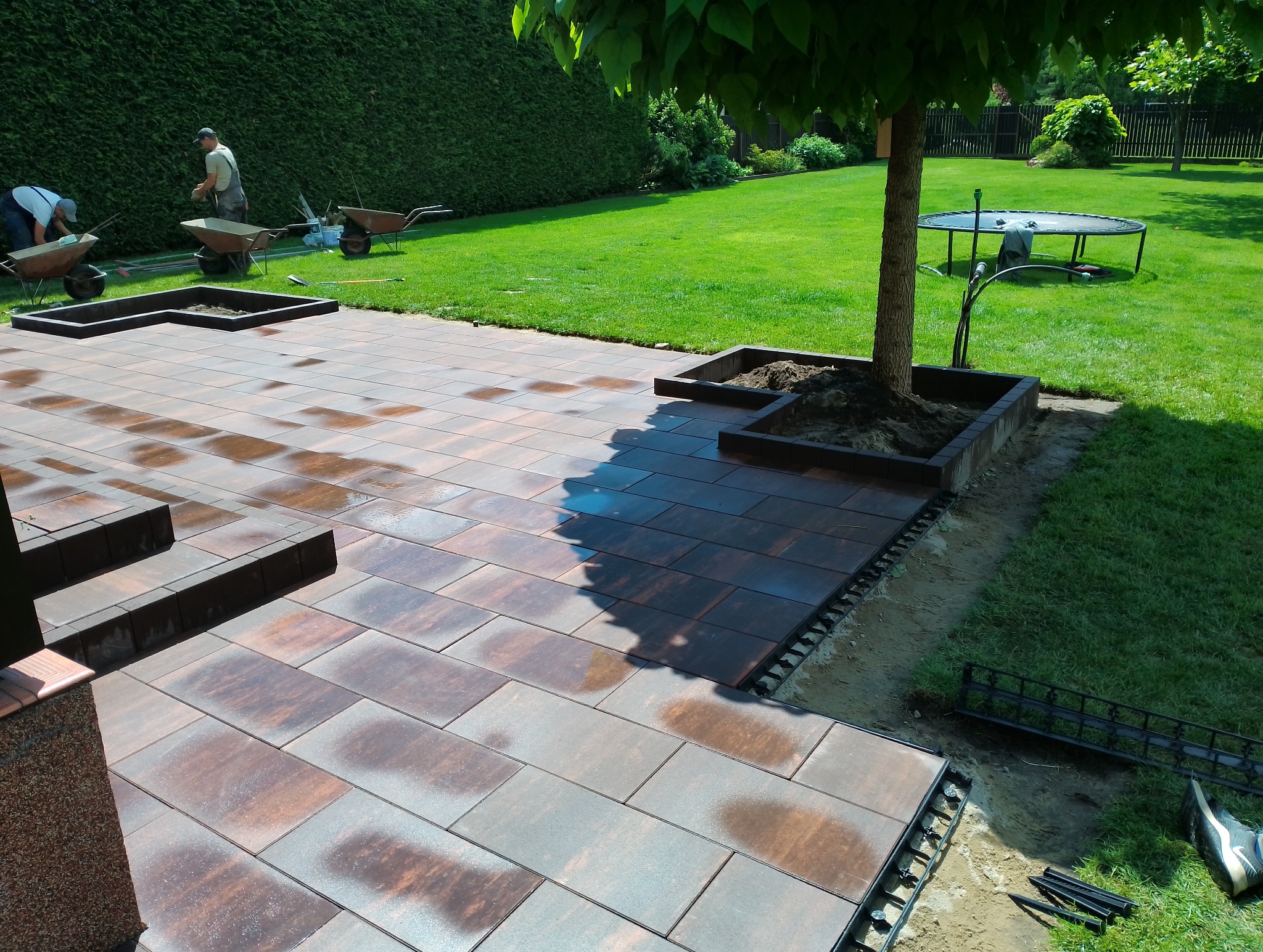 Układanie kostki brukowej w ogrodzie z wyznaczonymi miejscami na rośliny, widoczne elementy mocujące i pracownicy w tle.