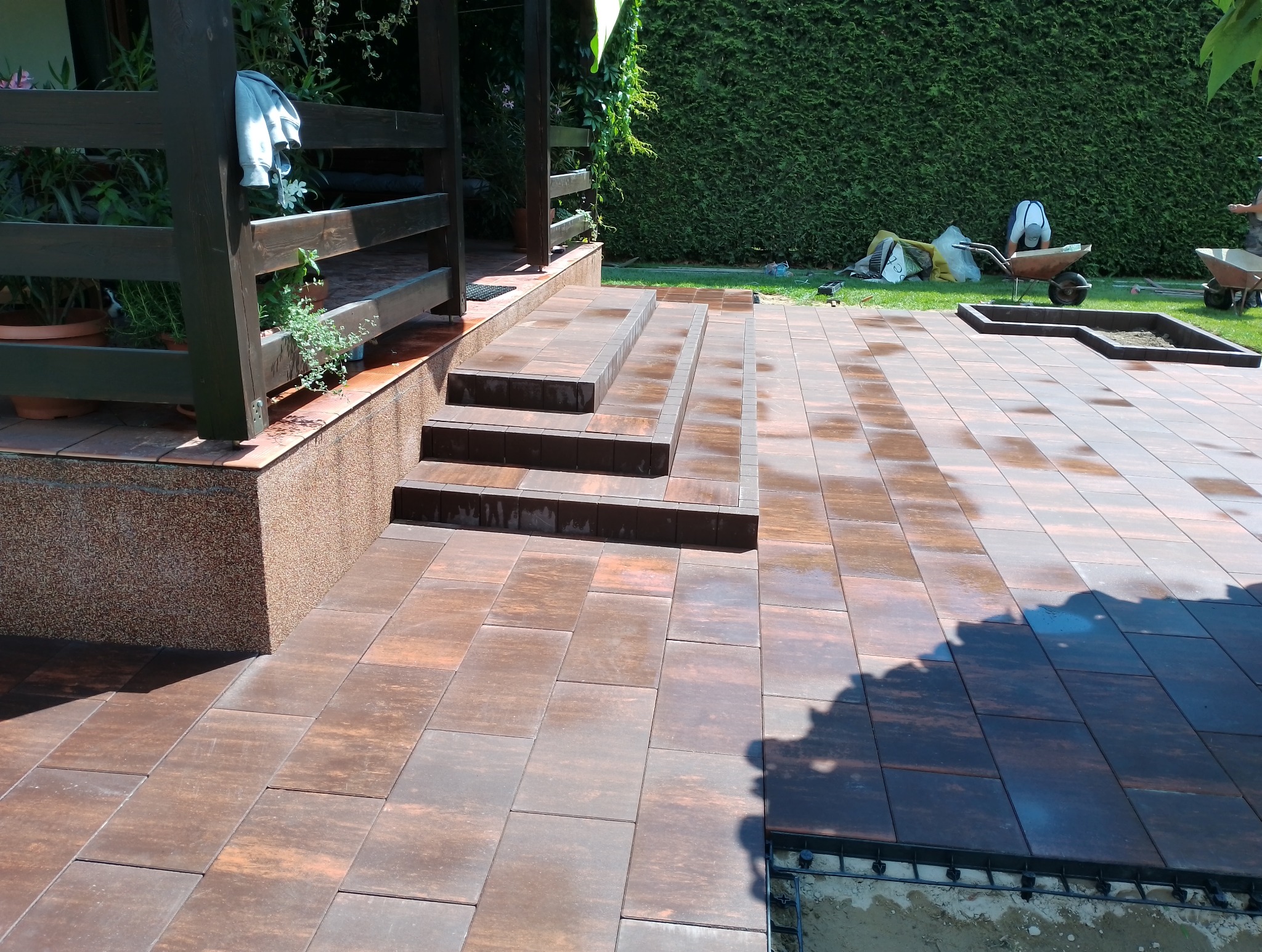 Ułożona, brązowo-pomarańczowa kostka brukowa prowadząca do schodów tarasu z drewnianą pergolą. W tle zielony żywopłot i prace ziemne przy budowie rabaty.