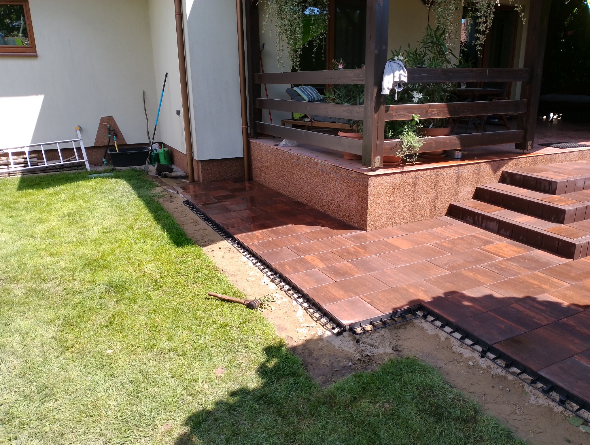 Brązowa kostka brukowa ułożona na tarasie domu jednorodzinnego, widoczne elementy mocujące przy trawniku, schody wejściowe i fragment ogrodu z narzędziami.