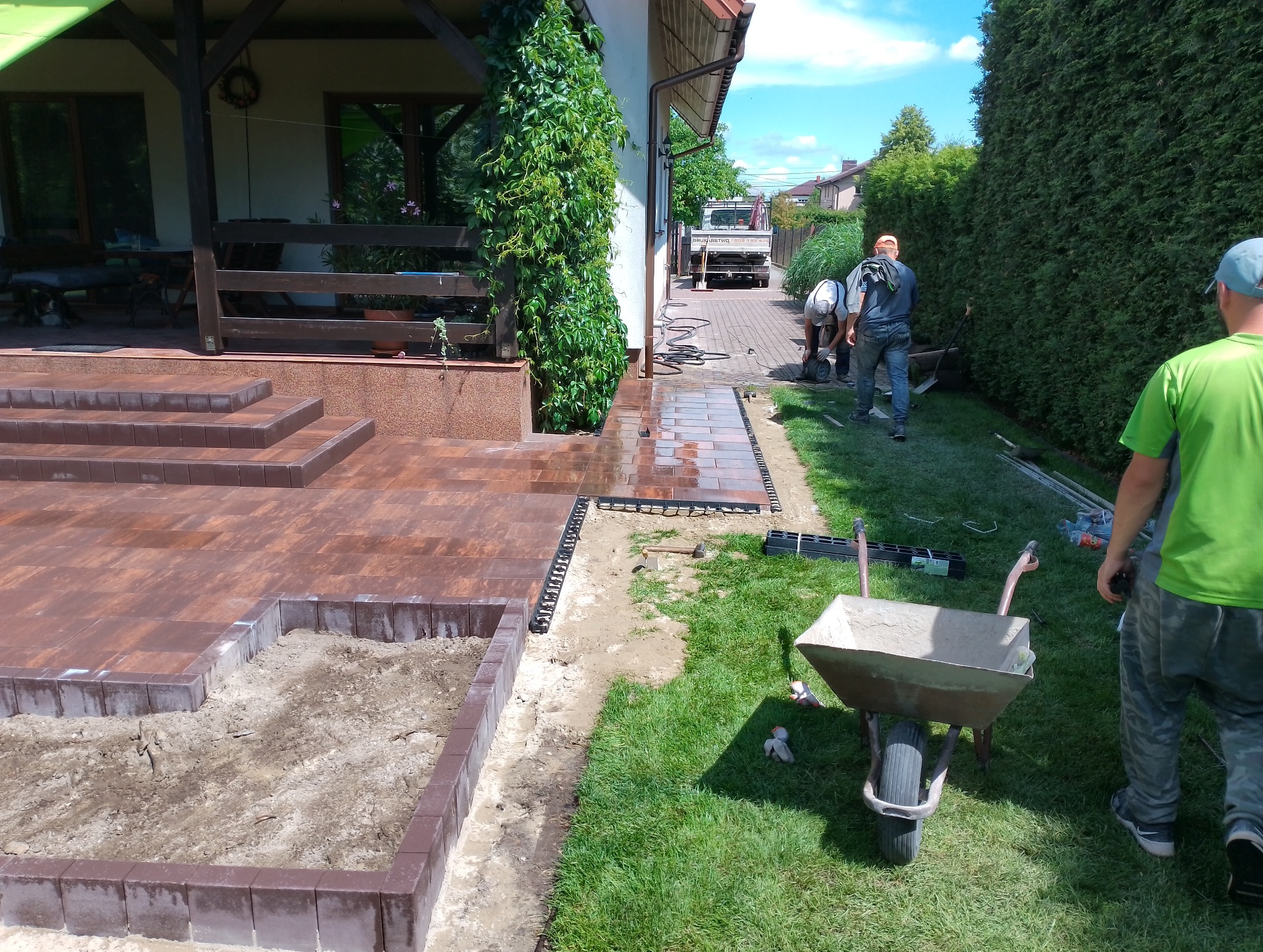 Montaż obrzeży trawnikowych przy nowej, brązowej kostce brukowej na posesji; w tle pracownicy układają kostkę na podjeździe, a przy budynku pnie się zielony bluszcz.