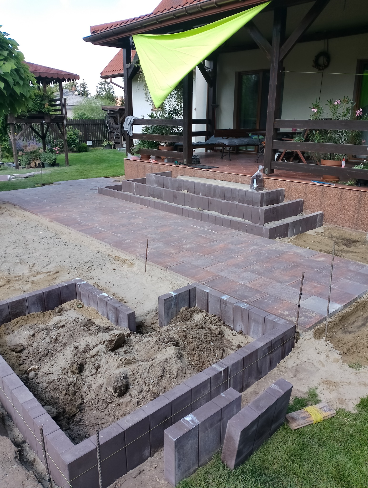 Układanie ścieżki z kostki brukowej w ogrodzie, widoczne obrzeża z kostki, częściowo ułożona nawierzchnia i przygotowany wykop pod kolejne elementy, w tle dom z tarasem.