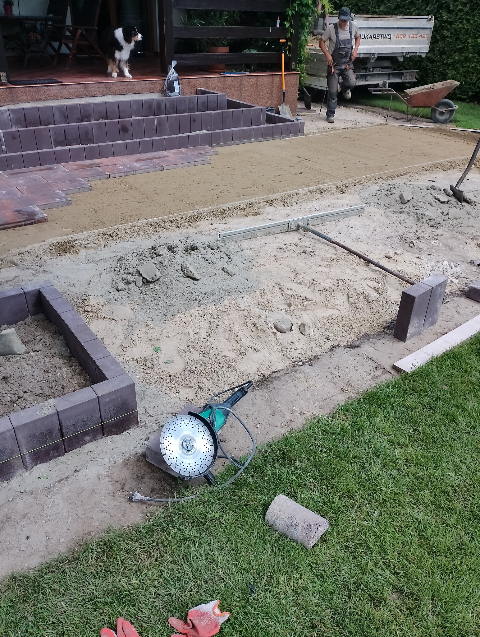 Układanie kostki brukowej w trakcie prac: widoczny fragment ułożonej kostki, przygotowane podłoże z piasku, ustawione obrzeża, poziomica, szlifierka kątowa na trawie oraz pracownik w tle.