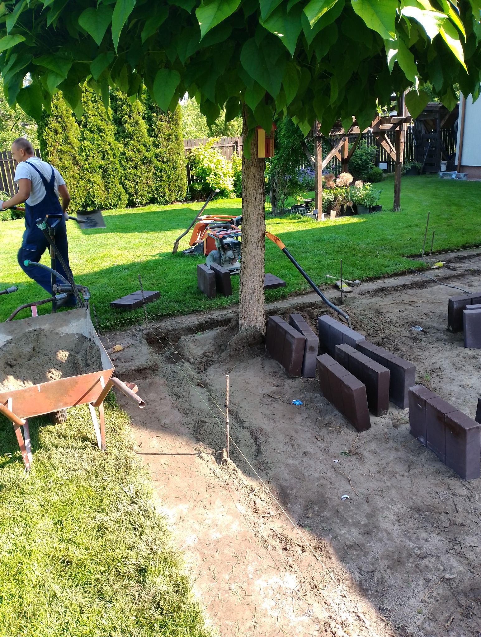 Pracownik w niebieskim kombinezonie z łopatą na tle wykopu pod ścieżkę z kostki, widoczny wózek z piaskiem, ubijarka i brązowe kostki brukowe.