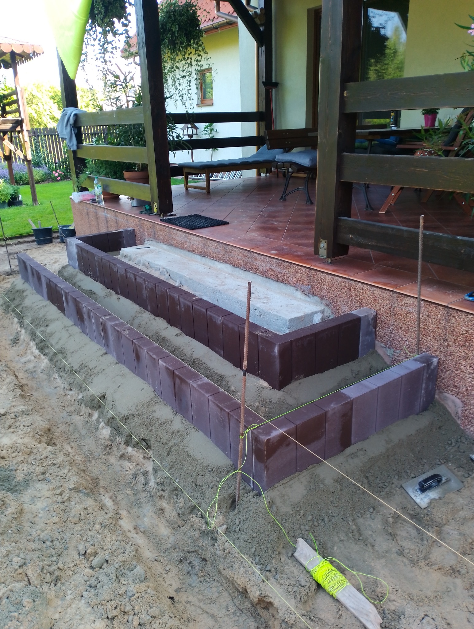 Montaż obrzeży z ciemnej kostki brukowej przy tarasie domu, widoczne sznurki wyznaczające linię, metalowe pręty, piasek i drewniany kołek z nawiniętym sznurkiem.