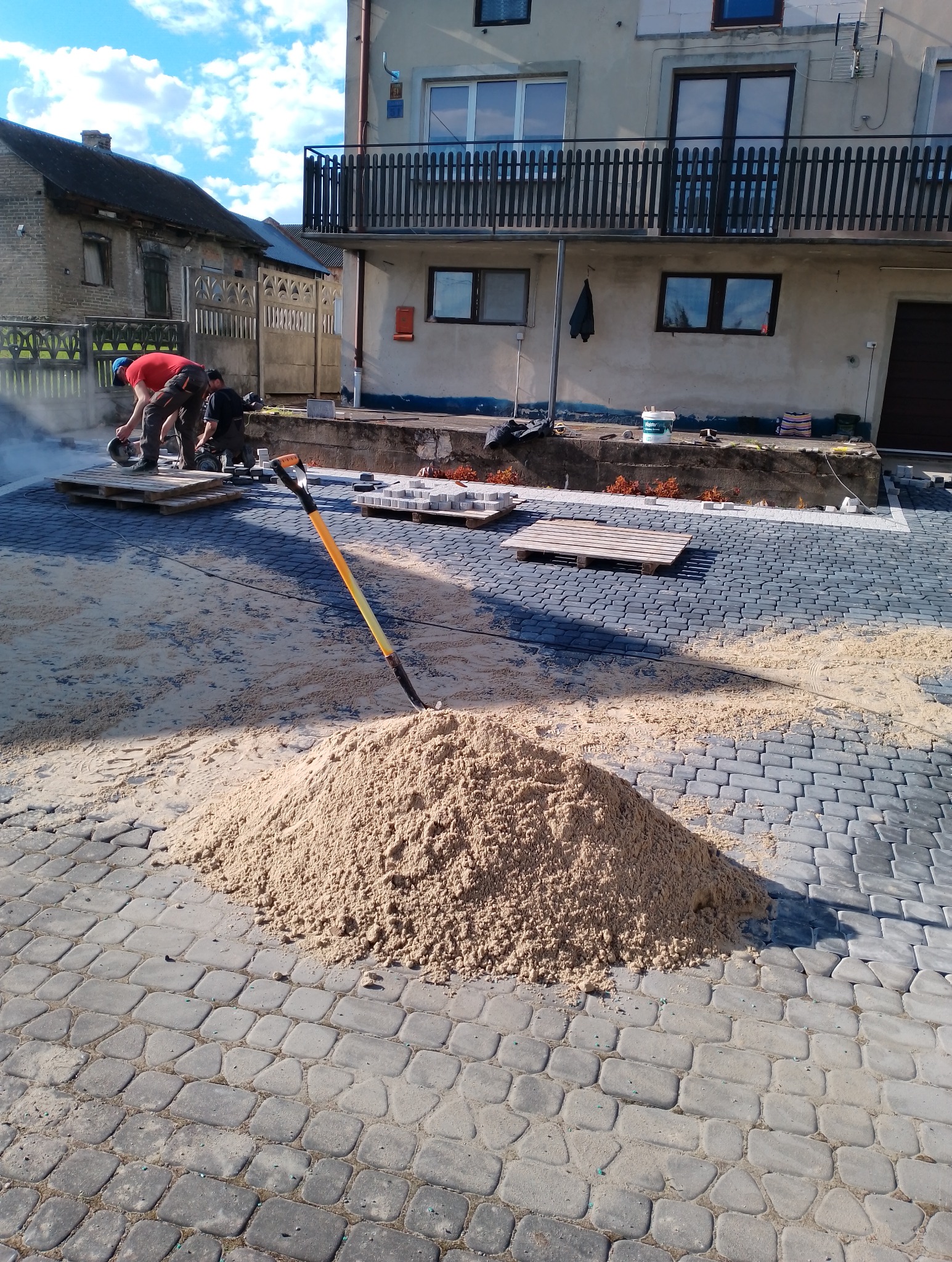 Dwóch pracowników tnie kostkę brukową na palecie w trakcie prac brukarskich, widoczny stos piasku i łopata z żółtym trzonkiem na tle częściowo ułożonej kostki.