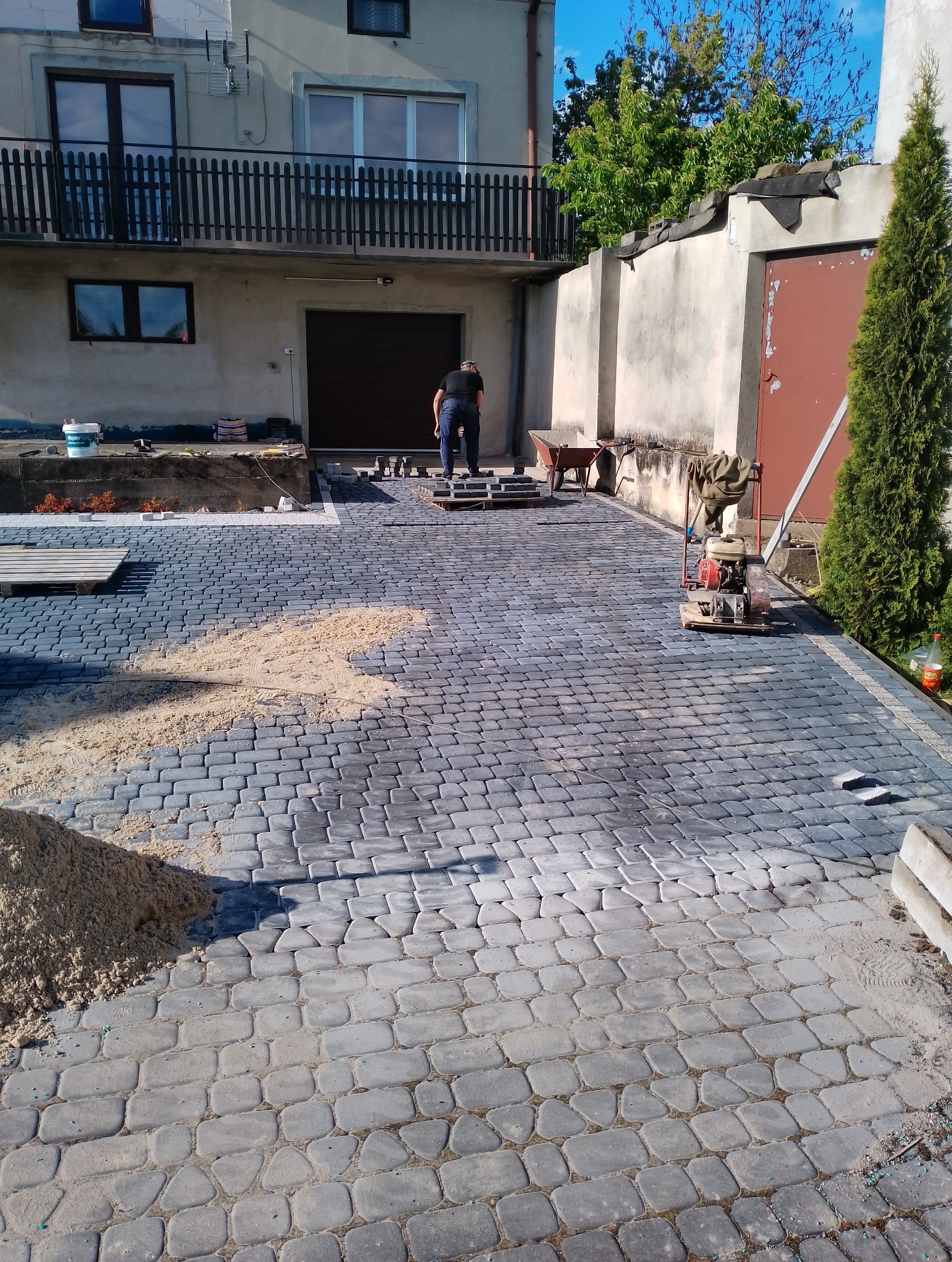 Ułożona z szarej kostki brukowej podjazd do domu z garażem, z widocznym pracownikiem, ubijarką i stertą piasku.