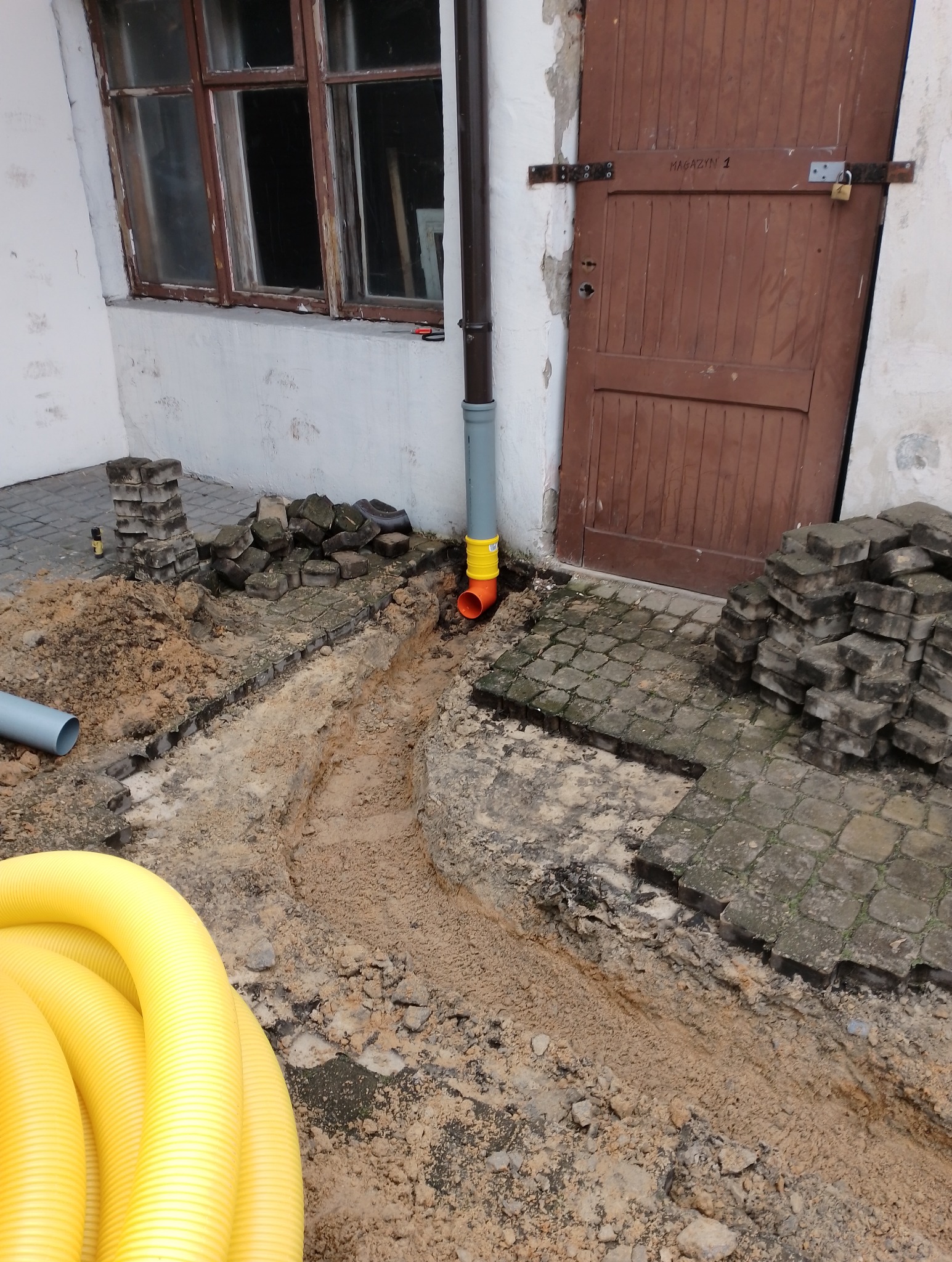 Przygotowanie podłoża pod kostkę brukową: wykop z podsypką piaskową, ułożone fragmenty kostki brukowej, rura drenażowa i stos kostki brukowej przy ścianie budynku z drewnianymi drzwiami 'Magazyn 1'.