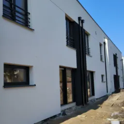 Nowoczesny budynek w trakcie wykańczania elewacji, widoczne fragmenty nieotynkowanych ścian z bloczków, biała, gładka elewacja, czarne obramowania okien i rur spustowych, teren budowy z resztkami...